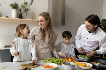 Joyful cute sibling kids helping parents to prepare dinner in home kitchen, cutting fresh vegetable at table, laughing, talking, chatting. Happy family couple teaching children to cookの写真素材