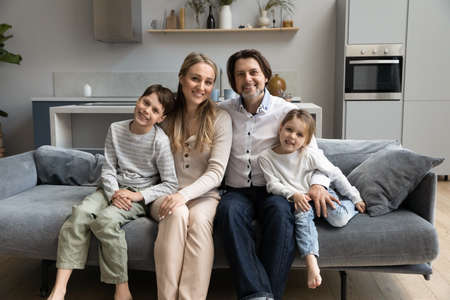Happy parents and two sibling children relaxing, hugging on sofa at home, looking at camera, smiling. Millennial family couple and kids posing on comfortable couch in new apartment front portraitの写真素材