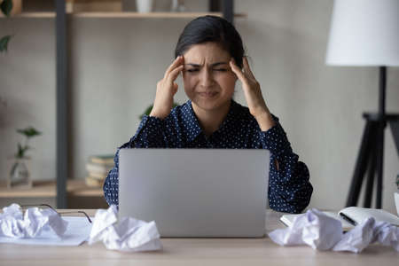 Unhappy annoyed Indian business woman feeling strong headache, anger, stress, nervous tension. Employee, office worker sitting at laptop and thrown papers, suffering from painful migraineの写真素材