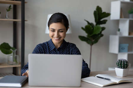 Happy Indian office worker typing on laptop computer at table, enjoying work, smiling. Female employee, student, businesswoman taking online training, reading email, chatting, making video callの写真素材