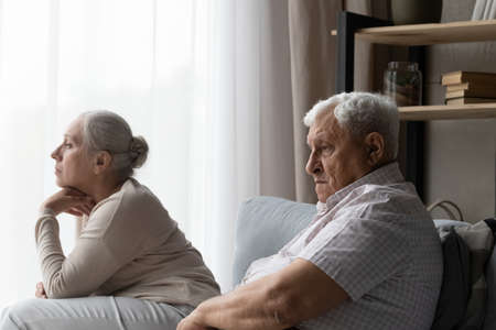 Frustrated unhappy old family couple sitting on sofa, looking in distance, experiencing grief, mourning together at home. Confused middle aged elderly 70s man and woman feeling depressed after quarrelの写真素材