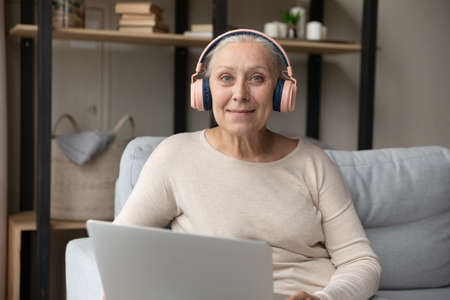 Portrait of smiling beautiful pleased mature retired hoary woman in headphones using computer applications, communicating distantly or e-learning at home, older people and modern technology usage.の写真素材