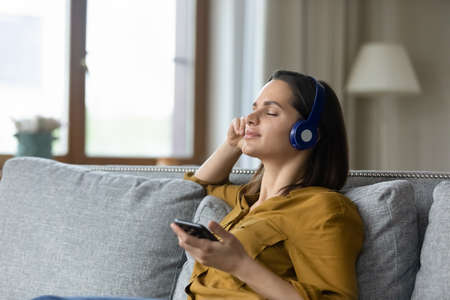 Happy pretty woman in wireless headphones enjoying ambient music on smartphone for recreation, reload, practicing stress relief meditation techniques, resting on comfortable sofa with closed eyesの写真素材