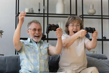 Happy sincere middle aged old retired family couple holding remote controllers, involved in playing online video games, enjoying entertaining virtual reality activity together at home, hobby pastime.の写真素材