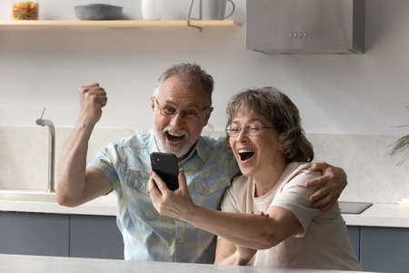 Overjoyed senior 60s family couple looking at cellphone screen, feeling excited getting message with amazing news, online lottery gambling auction win notification, celebrating internet success.の写真素材