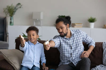 Happy funny African dad and son kid excited with videogame fighting in family virtual battle, playing online virtual video game on internet, using toy guns, aiming away at consoleの写真素材