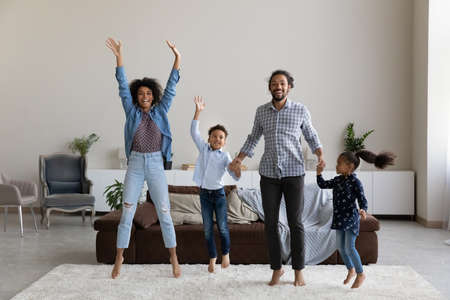 Excited cheerful African parents and little kids jumping on white soft carpet in living room, having fun, enjoying activity, leisure, home party, dancing to music, holding hands. Full length portraitの写真素材