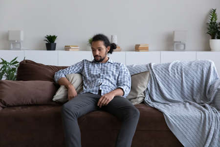 Passive bored sad African guy feeling tired, suffering from apathy, depression, sitting on sofa at home, looking away into vacancy, going through stress, loss. Frustration, fatigue conceptの写真素材