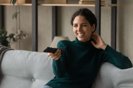 Joyful candid latina caucasian woman switching channels with remote controller, watching funny...
