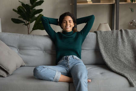 Happy young hispanic woman sitting on comfortable couch, feeling refreshed after day dream, meditating alone in living room. Mindful millennial lady relaxing with folded hands behind head at home.の写真素材