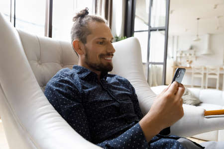 Smiling young hipster man looking at cellphone screen, enjoying playing games, web surfing...