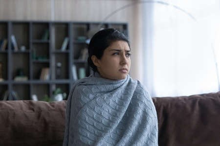 Stressed young Indian woman covered in warm plaid sitting on sofa at home, suffering from low temperature without heating system in living room or shivering having first flu symptoms.の写真素材
