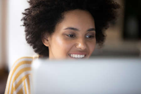Close up head shot smiling young African American female entrepreneur employee looking at computer screen, reading email with pleasant news, working distantly on online project at home or office.の写真素材