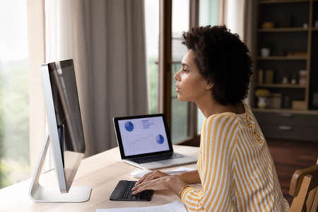 Focused young African American businesswoman looking at computer monitor, analyzing sales statistics data or marketing research results, typing preparing electronic document using different gadgets.の写真素材