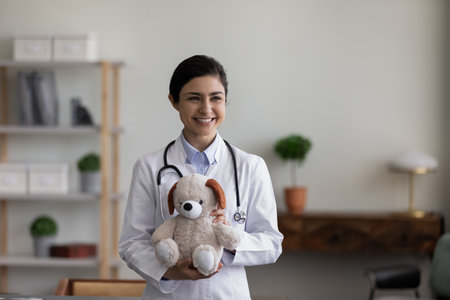 Smiling kind young Indian female pediatrician general practitioner doctor with teddy bear toy in hands, looking in distance waiting for small patients for checkup meeting in modern clinic office.の写真素材