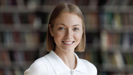 Happy pretty college student girl posing in university library with blurred bookshelves behind. Young millennial woman looking at camera with toothy smile head shot portraitの写真素材