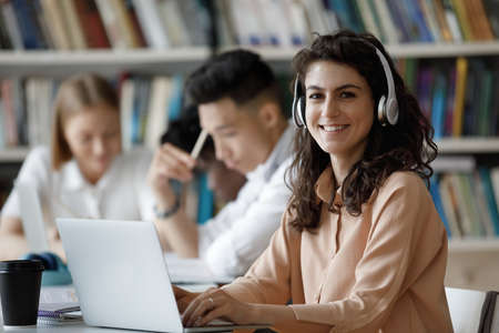 Happy gen Z Latin student girl in headphones working on research study project in library, watching online presentation on laptop, learning webinar, sitting by classmates. Head shot portraitの写真素材