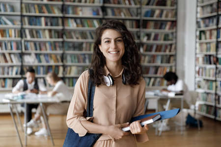 Happy Hispanic gen Z student girl with headphones visiting public library for work on study research project, holding learning papers, notebook, looking at camera, smiling. Head shot portraitの写真素材