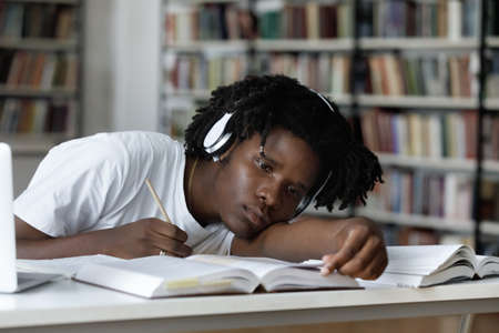 Tired bored lazy Black student listening to audio lesson, webinar, writing notes, reading open book in library with head rested on table. Overworked exhausted college guy working on study projectの写真素材
