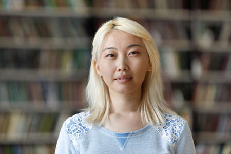 Pensive pretty Asian blonde student girl posing in university library with blurred background behind. Young Chinese hipster woman with pale hair looking at camera head shot portraitの写真素材