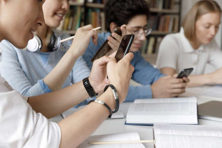 Group of young gen Z students browsing Internet in campus library, using smartphones at work table with books, collaborating on project for class, chatting online. Close up of hands holding phoneの写真素材