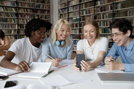 Happy cheerful multiethnic group of students sharing mobile phone, making video call in public college library, watching learning webinar, online lesson, consulting teacher, smilingの写真素材
