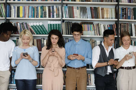 Multiethnic group of freshman students engaged in cellphone using, standing in row in library at bookshelf, chatting online, browsing internet, scrolling social media, suffering from addictionの写真素材