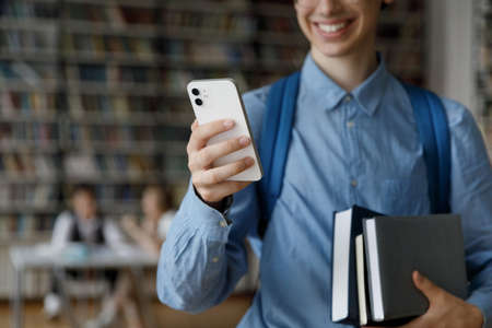 Happy young gen Z student guy using online app, internet service on smartphone, holding stacked books, textbooks, smiling, visiting college library. Hand holding mobile phone close upの写真素材