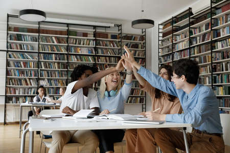 Interracial group of college girl and guys giving group high five gesture over table in university...