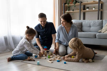 Happy bonding young couple parents and joyful little preschool children son daughter playing toys, sitting on floor carpet, enjoying creative playtime hobby activity together in modern living room.の写真素材