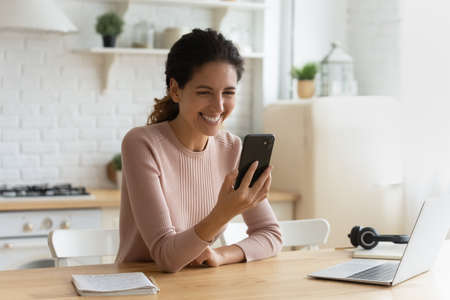 Young 30s smiling Hispanic woman sit at table in home kitchen holds modern smartphone laughing at video, enjoy videocall event, funny internet content, on-line fun, amusing mobile application conceptの写真素材