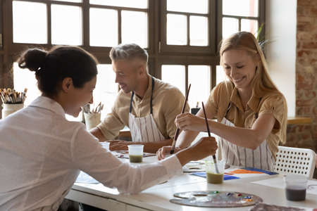 Cheerful diverse art school classmates girls talking on class, drawing in paints at table, chatting,...