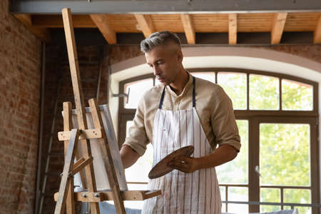 Serious busy professional artist man in craft apron working in studio, painting on canvas, using...