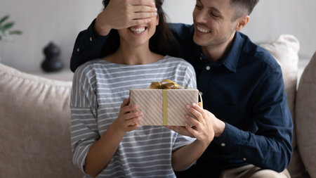 Crop close up loving man boyfriend preparing surprise for woman, covering her eyes with hand, smiling curious girlfriend holding present box, couple celebrating anniversary or birthday togetherの写真素材