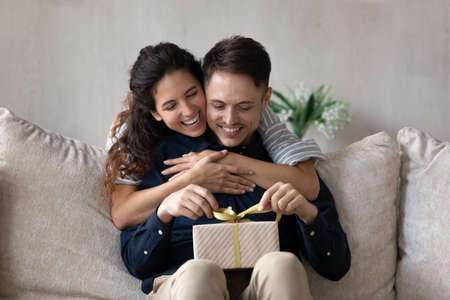 Loving girlfriend congratulating boyfriend, hugging from back, excited curious man holding gift box,...
