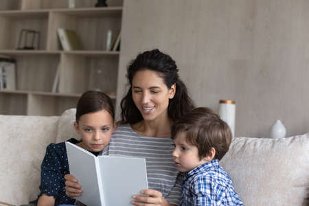 Smiling Hispanic mother reading book to little son and daughter sitting relaxing on cozy couch at home, caring young mom telling to kids funny fairy tale story, spending leisure time weekend togetherの写真素材