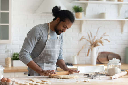 Happy handsome young 30s African American man in apron rolling out dough for homemade pastry, enjoying preparing biscuit cookies in modern light kitchen, cooking hobby activity pastime concept.の写真素材