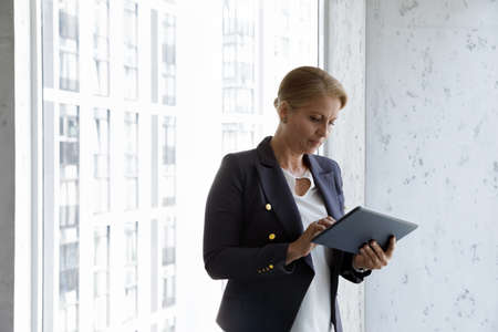 Serious confident mature businesswoman using online app on tablet computer in office hallway at window, watching virtual video presentation, reading report, email letter. Business communicationの写真素材