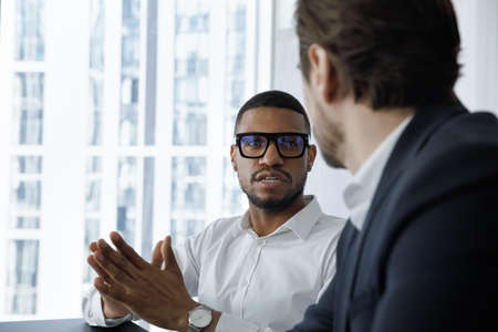 Serious engaged African employee in glasses talking to coworker, business partner, speaking, telling about project. Multiethnic workers, professionals discussing work tasks at workplaceの写真素材