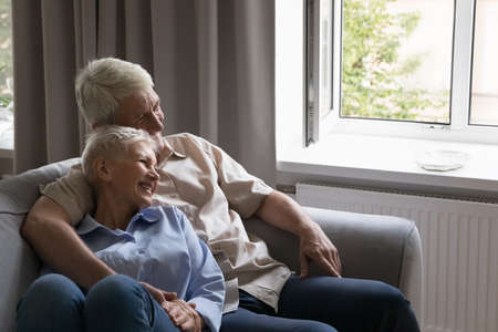 Happy older couple in love relaxing on home couch at open window. Senior husband hugging mature wife with care, support, tenderness, resting on sofa, talking, discussing future plans, laughingの写真素材