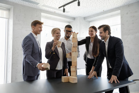 Happy successful diverse business team of different ages building blocked tower on office table together, having fun, laughing, working on teambuilding, unity, cooperation skills, celebrating successの写真素材