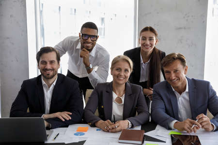 Happy diverse different aged business team sitting, standing at meeting table in office together, looking at camera, smiling. Group head shot portrait of successful young and mature coworkersの写真素材