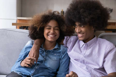 Cheerful happy Black couple of hipster teenagers, students enjoying dating, leisure time together at home, relaxing sitting on couch, talking, chatting, laughing. Youth, love conceptの写真素材