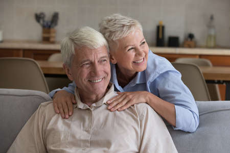 Cheerful middle age senior married couple relaxing in home living room. Mature wife hugging husband...