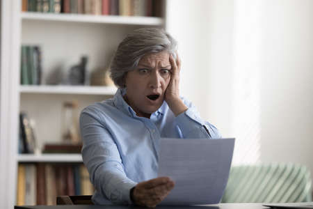 Shocked stressed middle aged mature 50s woman looking at paper document, feeling frustrated reading...