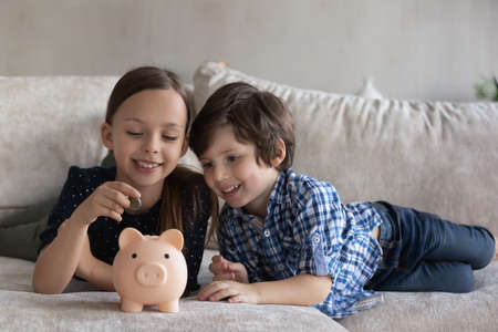 Cheerful sibling little kids putting coin into pink piggybank, playing financial game, resting on couch at home. Children saving money, cash, learning economy, planning future investment.の写真素材