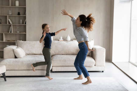 Excited active mom and happy daughter kid having fun, going wild, dancing to music in living room, jumping on floor, practicing exercises, moving bodies, listening to radio. Home activity conceptの写真素材