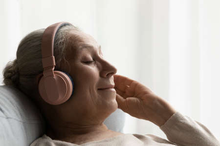 Happy calm relaxed older woman in big headphones resting on comfortable couch, listening to music for relaxation, meditation, enjoying break, leisure time at home, smiling with closed eyesの写真素材