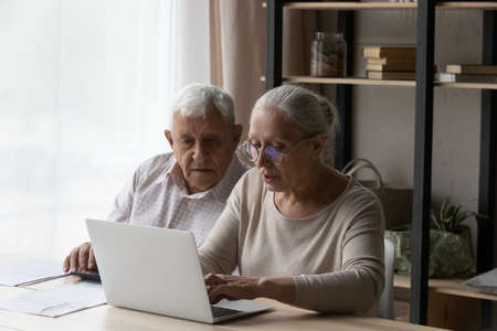 Senior married couple paying taxes, domestic bills, insurance fees on Internet. Husband and wife managing budget, counting expenses, savings, using laptop computer at table togetherの写真素材