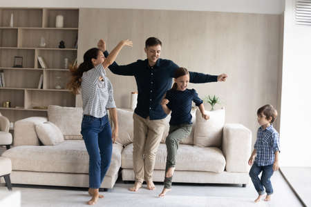 Joyful family of new homeowners dancing in living room, celebrating relocation at home. Happy young parents and two active kids having fun together, jumping to music, exercising, enjoying partyの写真素材